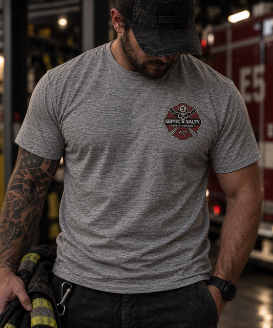 Male firefighter wearing a heather gray Firehouse Recliner Rodeo T-shirt with the Septic & Salty Supply Company Maltese cross logo on the front left chest inside a fire station.