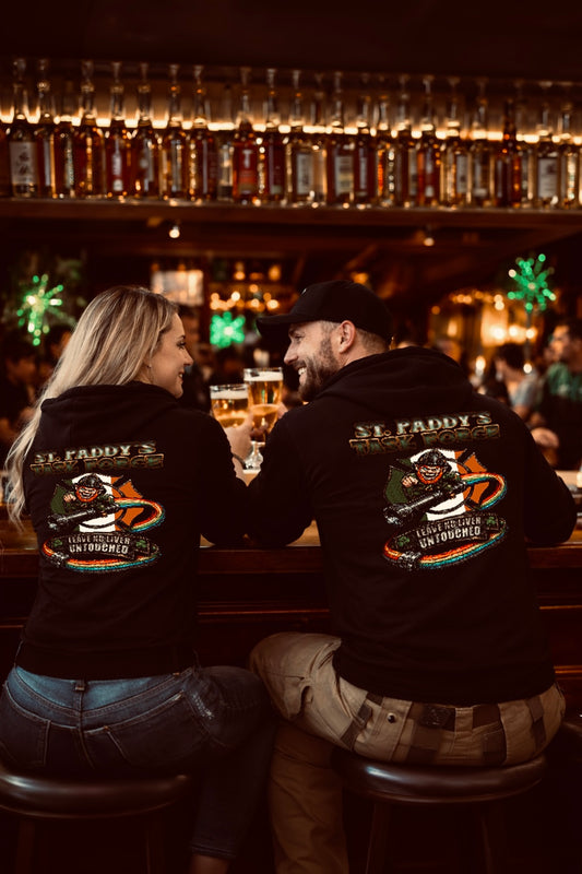 Man and woman wearing black St. Paddy’s Task Force hoodies at a bar, displaying the back graphic with firefighter leprechaun and rainbow hose design.