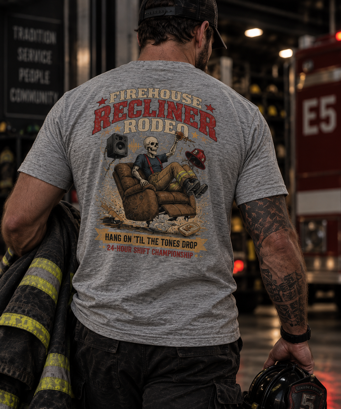 Rear view of a firefighter wearing the heather gray Firehouse Recliner Rodeo T-shirt featuring the large skeleton firefighter recliner graphic inside a firehouse apparatus bay.