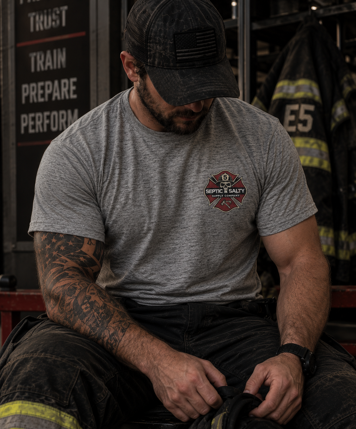 Male firefighter seated in a firehouse wearing a heather gray Firehouse Recliner Rodeo T-shirt with the Septic & Salty Supply Company Maltese cross logo on the front left chest.