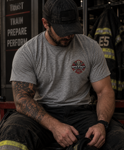 Male firefighter seated in a firehouse wearing a heather gray Firehouse Recliner Rodeo T-shirt with the Septic & Salty Supply Company Maltese cross logo on the front left chest.