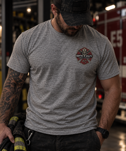 Male firefighter wearing a heather gray Firehouse Recliner Rodeo T-shirt with the Septic & Salty Supply Company Maltese cross logo on the front left chest inside a fire station.