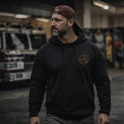 Firefighter wearing the black "Firehouse Fistfights" hoodie with left chest Septic & Salty Supply Co. logo — lifestyle shot in a firehouse bay with apparatus visible in the background.