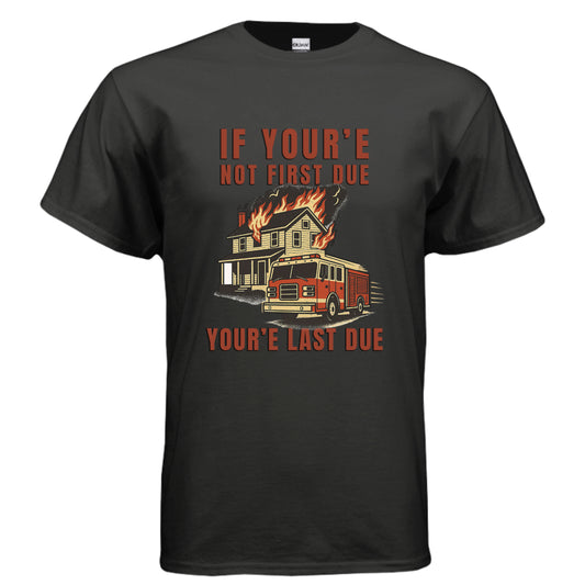 Cartoon-style firefighter shirt design showing a red fire engine in front of a burning house with the caption “If You’re Not First Due, You’re Last Due,” capturing firefighter competition and dark station humor.