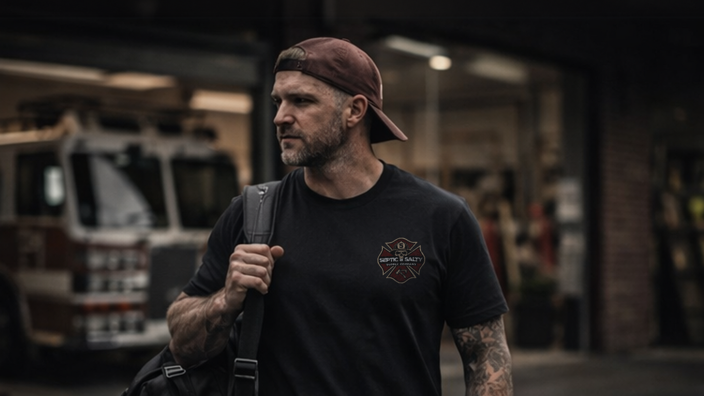 Man wearing a black Septic & Salty Supply Company t-shirt with left-chest Maltese cross logo, carrying a gear bag outside a fire station with an engine visible in the background​​​​​​​​​​​​​​​​