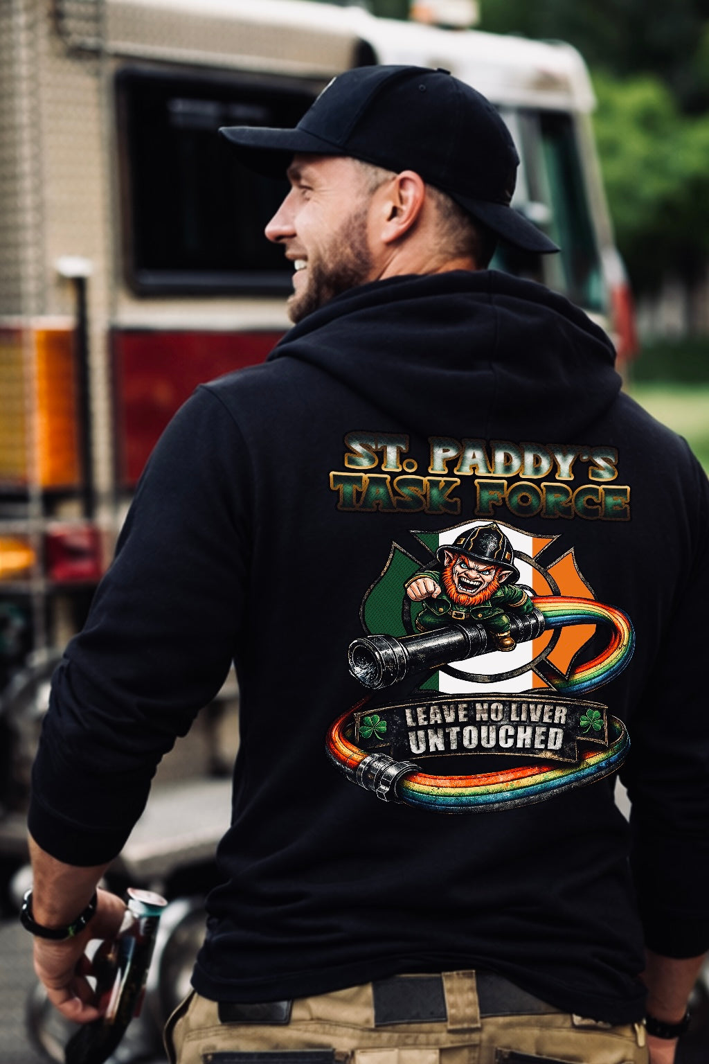 Firefighter standing near a fire truck wearing a black St. Paddy’s Task Force hoodie with large back graphic featuring Irish-themed firefighter artwork and slogan.